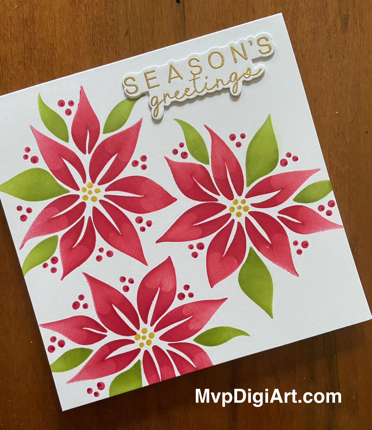 A handmade square Christmas card with a gold 'Season's Greetings' text and a poinsettia pattern on the front, including red flowers, green leaves, and red berries.