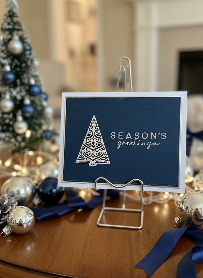 Navy card with silver tree design and 'Season's greetings' text on a stand, Christmas decorations in the background.