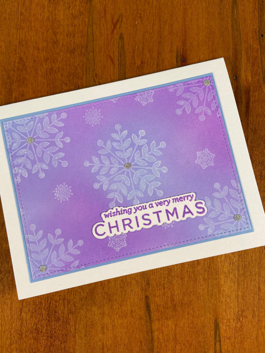 Handmade Christmas card with blue and purple ink blended background with a white snowflakes design with silver glitter accents on white notecard; text ‘wishing you a merry Christmas’