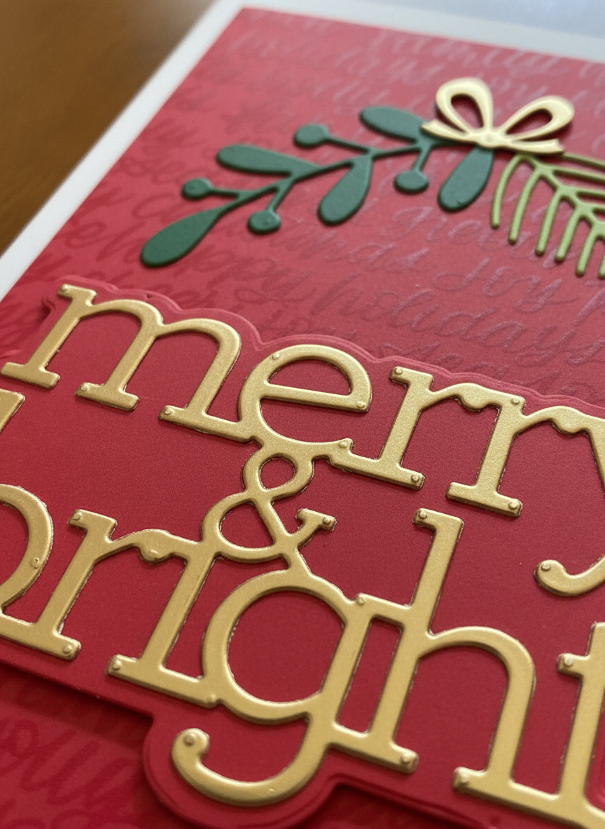 Close-up detail of Christmas card showing die-cut spruce and leaves tied with lustrous gold bow and 'merry & bright' golden text on red background