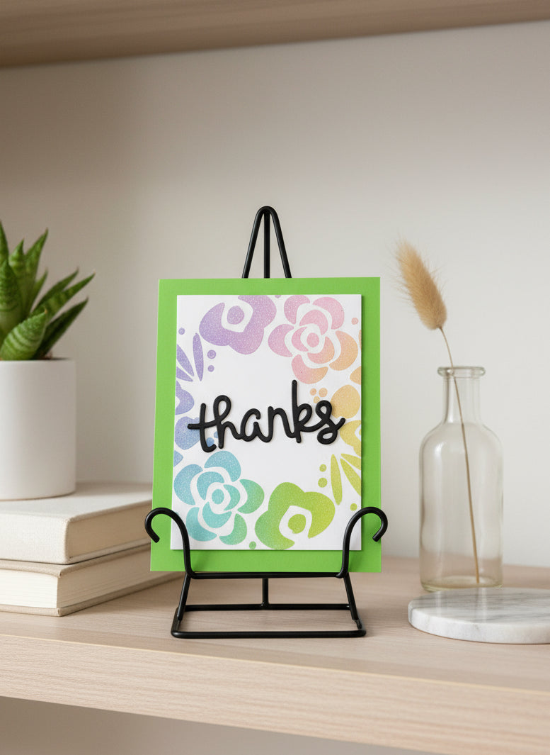 Handmade thank you card with pastel floral wreath on green notecard displayed in stand on decorated shelf with books and plant