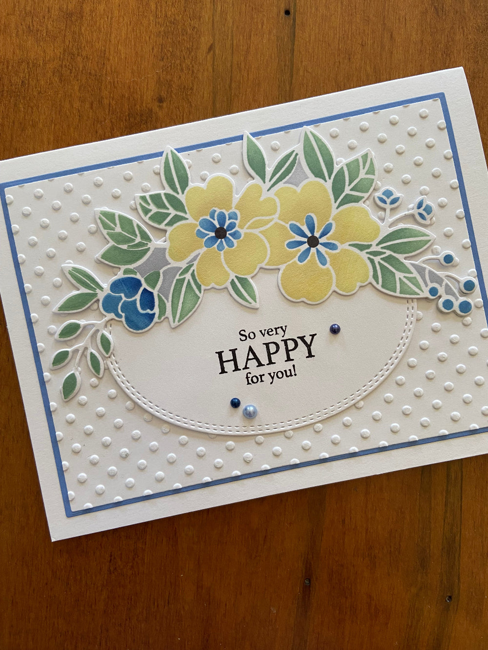 A handmade wedding card with a floral banner featuring die-cut flowers in yellows, blues, and greens, and the text 'So Happy For You' stamped on it. 
