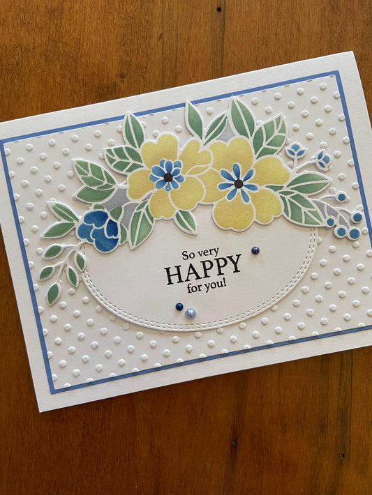 A handmade wedding card with a floral banner featuring die-cut flowers in yellows, blues, and greens, and the text 'So Happy For You' stamped on it. 