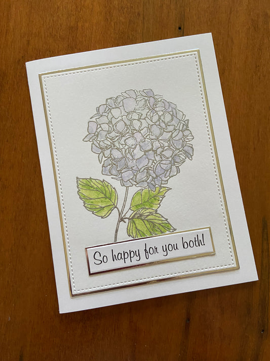Handmade wedding card featuring silver-embossed hydrangea clusters hand-watercoloured in purple and green, with a "so happy for you both" sentiment strip on shimmering silver cardstock against a white notecard.