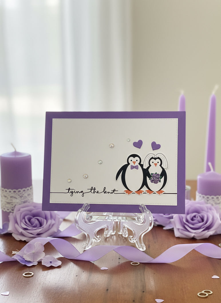 Handmade wedding card with penguin bride and groom, purple hearts, and pearl accents displayed on decorated table