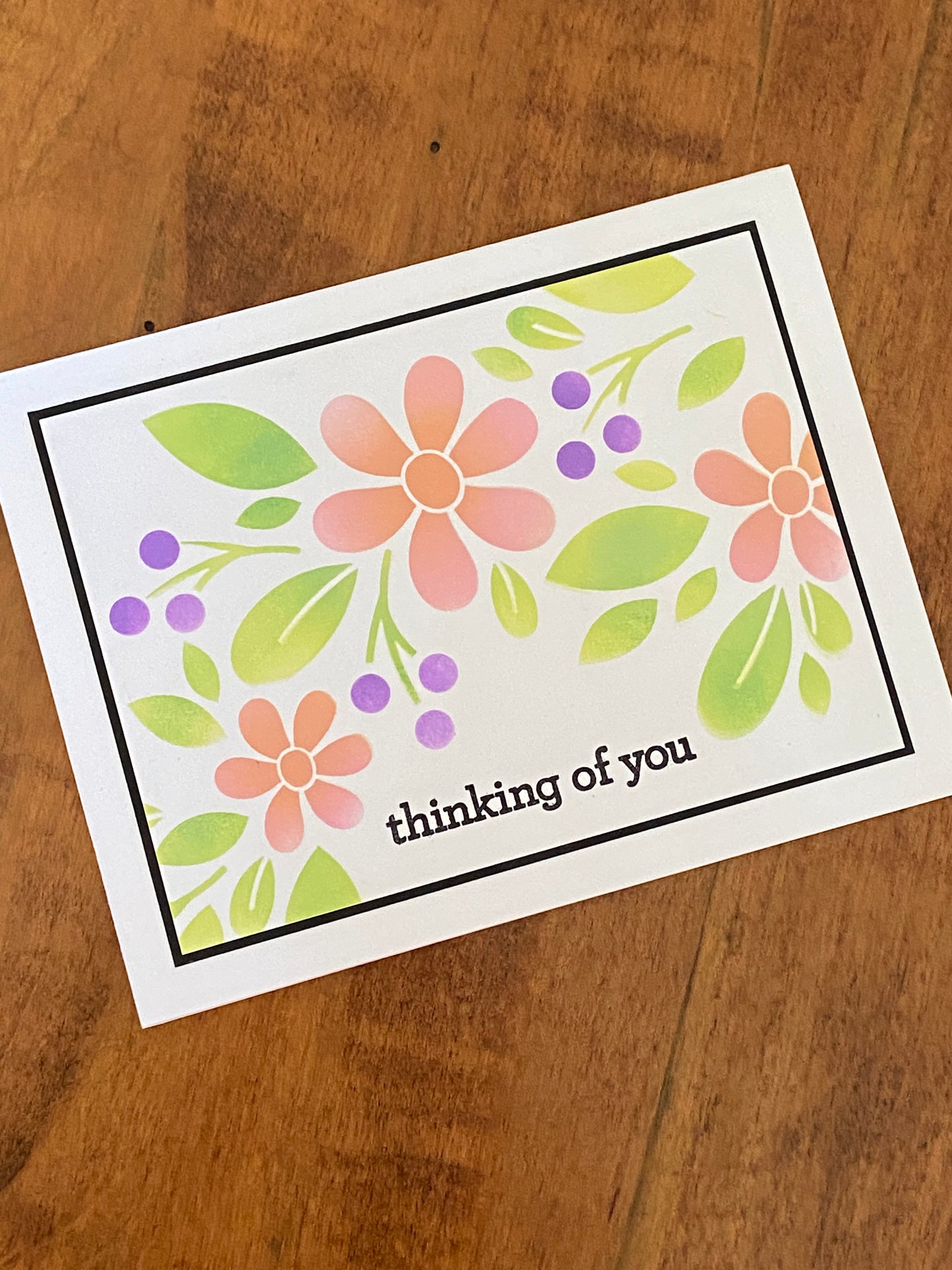 A coral daisy like flower, with green foliage and purple berries are hand stencilled onto a white background.  A thin black matte surrounds the panel which is mounted onto a white notecard.   A simple stamped "thinking of you" sentiment is stamped in black below the flowers and leaves.