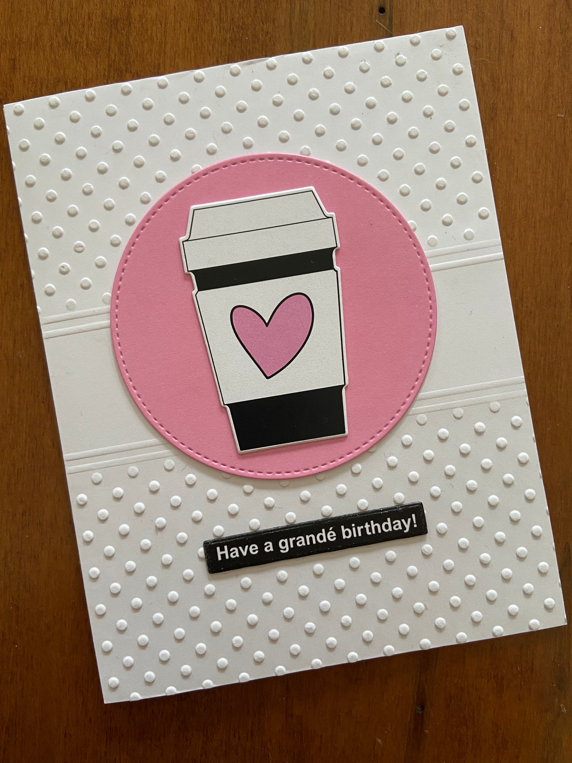 The card front is a Swiss Dot embossed background. A pink circle die cut is centred on the card with a cute little to go coffee cup adorned with a wonky pink heart. A simple black sentiment strip with white print says "Have a grandé birthday!"