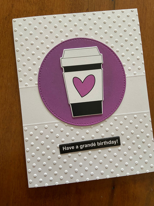 The card front is a Swiss Dot embossed background. A purple circle die cut is centred on the card with a cute little to go coffee cup adorned with a wonky purple heart.  A simple black sentiment strip with white print says "Have a grandé  birthday!"