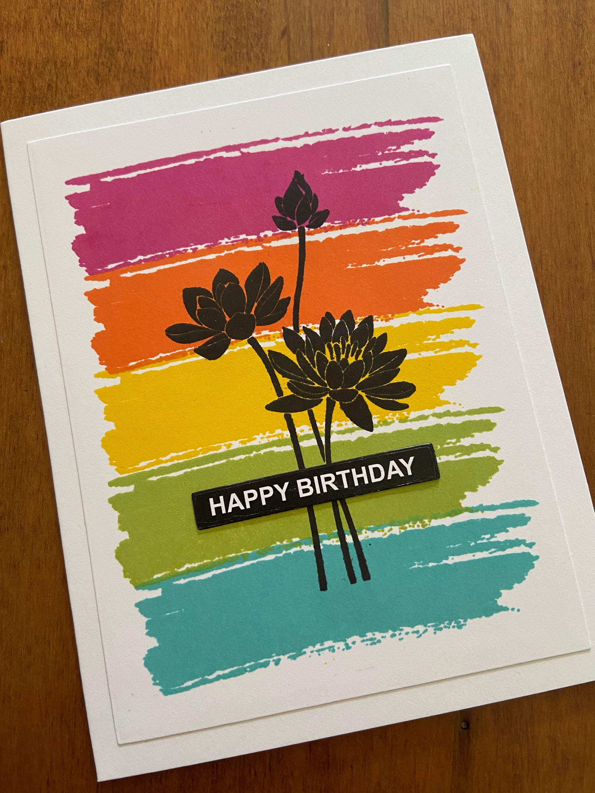 Dark pink, orange, yellow, lime green and turquoise paint stroke stamps background. A lotus silhouette stamp over top and a happy birthday sentiment over top. 