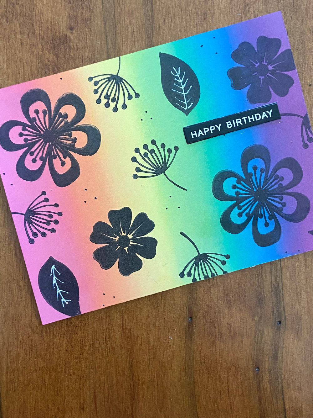 A handmade birthday card with a rainbow ink background and floral silhouettes. The card has 'Happy Birthday' text and a space inside for a personal message.