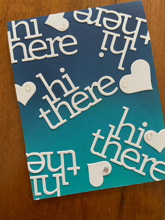 A greeting card with a combination of blue and teal inks, featuring white text that reads 'hi there' and decorative white hearts.