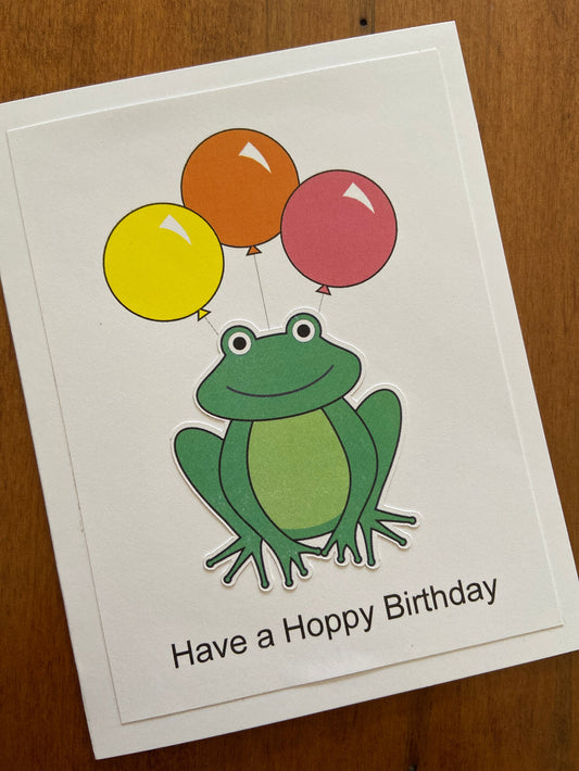 A birthday card with a digital printed green frog holding colourful balloons, with the text 'Have a Hoppy Birthday' on the front.
