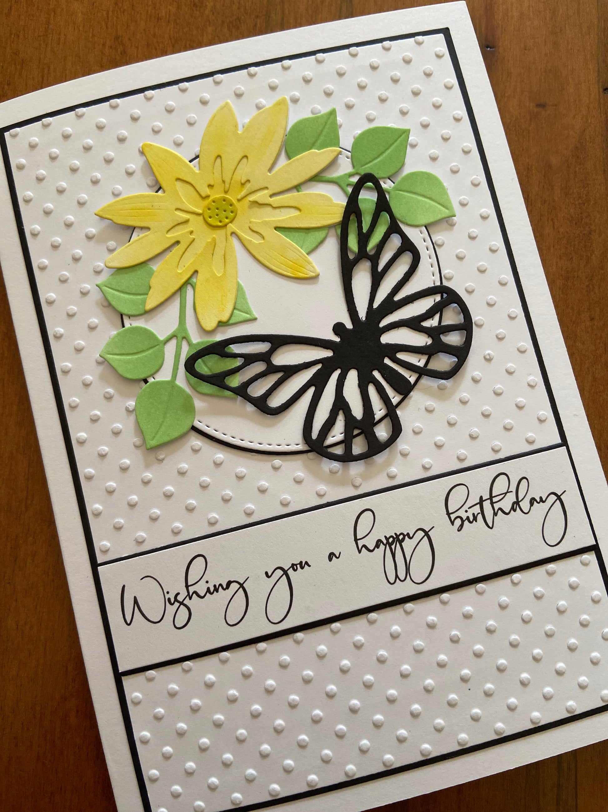 A 5 x 7 birthday card that is embossed with a swiss dot pattern background, with a yellow daisy and green foliage focal image on top. A black die cut butterfly is visiting the daisy. A scripty  wishing you a happy birthday sentiment completes the card.