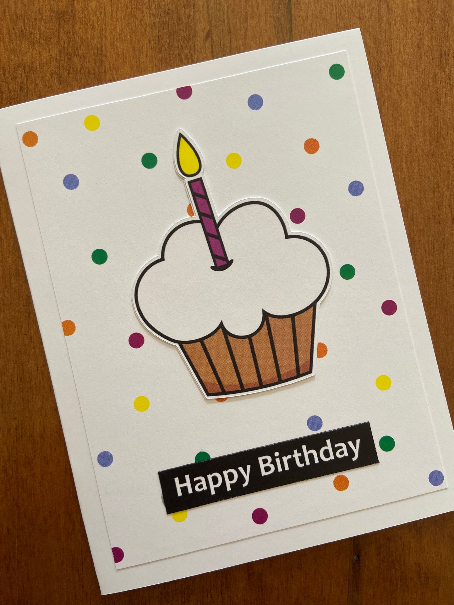 Colourful polka dots on a white background with a popped up cupcake and a single candle die cut. This card is completed with a Happy Birthday sentiment strip. below the cupcake.